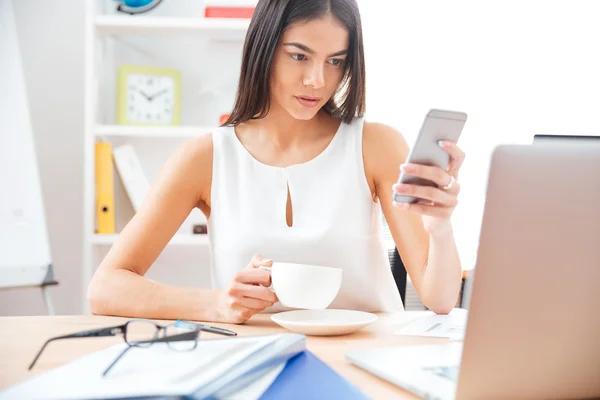 Empresária segurando xícara com café e usando smartphone — Fotografia de Stock