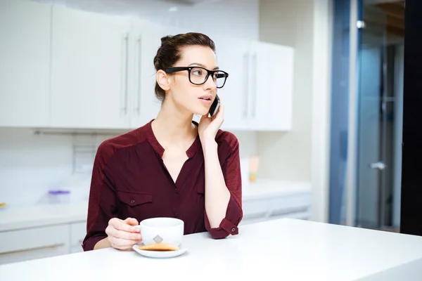Woman in glasses drinking tea and using smartphone on kitchen — Stock Photo, Image
