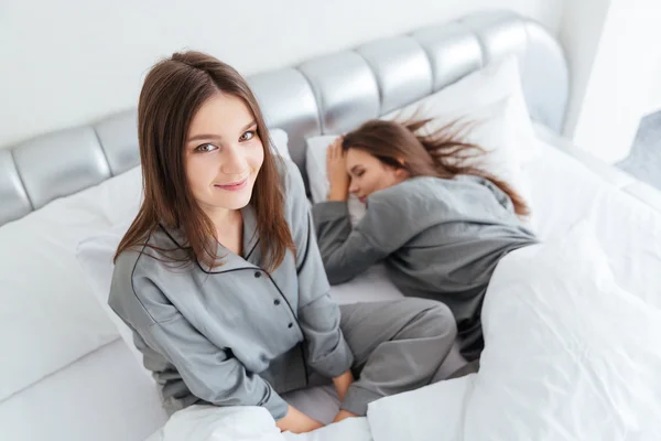 Cheerful woman sitting on bed near her sleeping sister twin