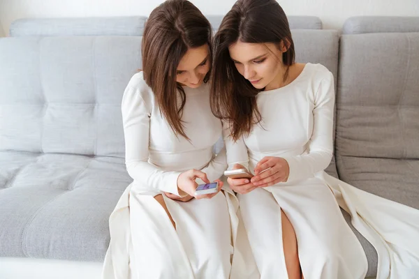 Dvě sestry dvojčata sedící na gauči a používání mobilních telefonů — Stock fotografie