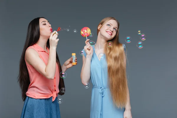 Dos mujeres hermosas alegres comiendo piruleta colorida y burbujas que soplan — Foto de Stock