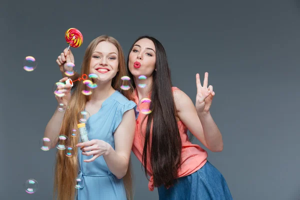 Dos mujeres jovenes juguetonas felices con burbujas que soplan coloridas de la piruleta — Foto de Stock