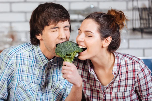 Casal mordendo brócolis juntos na cozinha — Fotografia de Stock