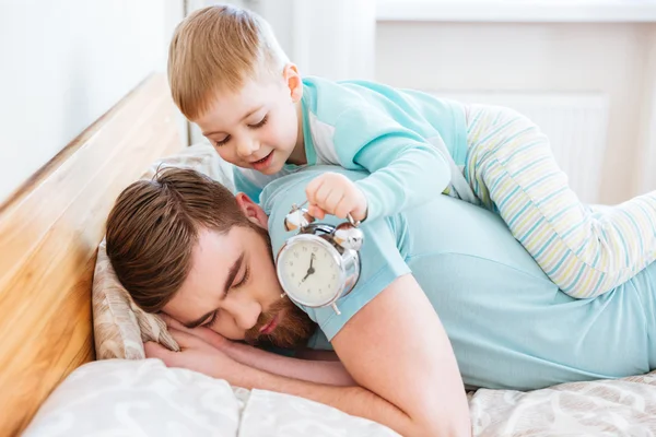 Pequeño hijo sosteniendo el despertador cerca de dormir oído padre — Foto de Stock