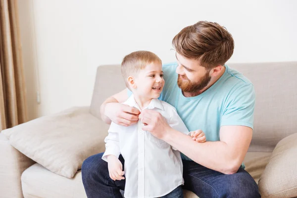 Счастливый отец сидит на диване и одевает своего маленького сына — стоковое фото