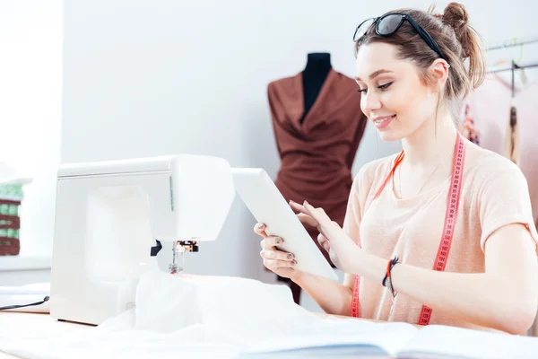 Mujer sonriente costurera sentada y usando tableta en el trabajo —  Fotos de Stock