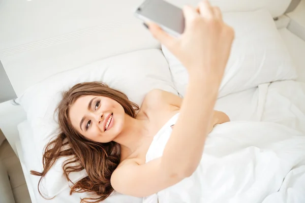 Mujer sonriente acostada en la cama y haciendo foto selfie —  Fotos de Stock