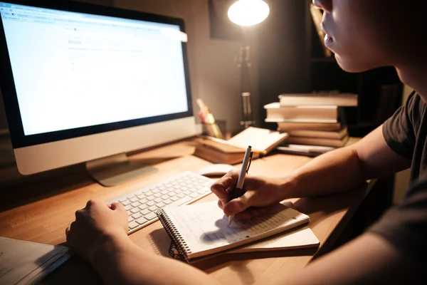 Homem escrevendo no bloco de notas e usando o computador no quarto escuro — Fotografia de Stock