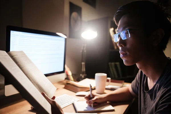 Hombre leyendo libro y escribiendo en bloc de notas sentado cerca de la computadora —  Fotos de Stock