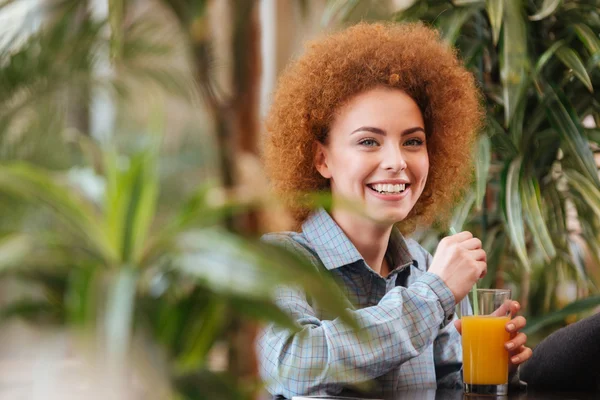 Mulher alegre bebendo suco de laranja no café com plantas verdes — Fotografia de Stock