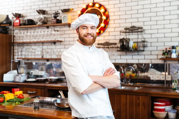 Cozinheiro chef sorridente de pé com as mãos dobradas na cozinha — Fotografia de Stock