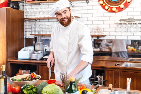 Cheerful jovem cozinheiro cortar carne e fazer salada de legumes — Fotografia de Stock