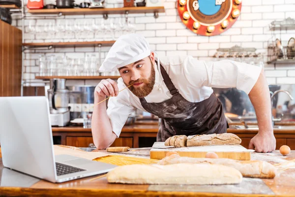 Pão de corte pensativo padeiro e usando laptop na cozinha — Fotografia de Stock