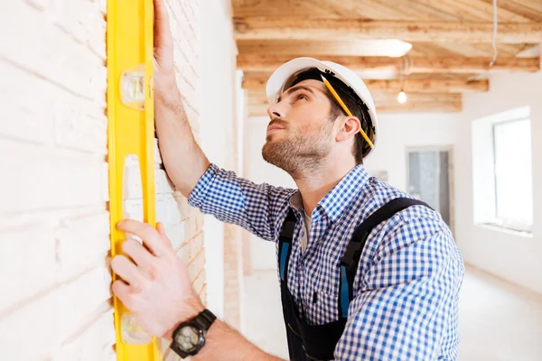 Construtor em uniforme segurando um nível contra a parede dentro de casa — Fotografia de Stock