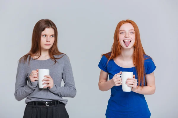 Dos felices bromas y tristes mujeres jóvenes frunciendo el ceño bebiendo té — Foto de Stock