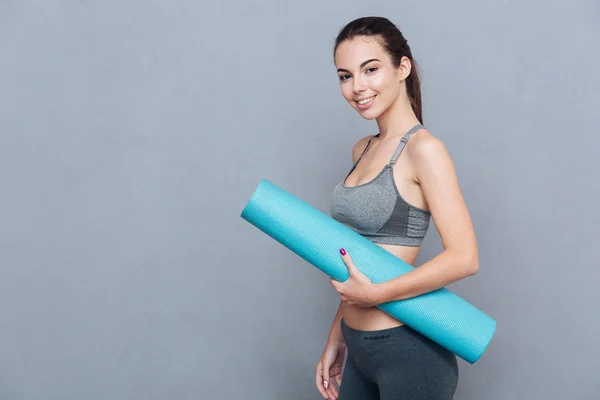 Attractive cheerful sportswoman holding yoga mat isolated over grey background — Stock Photo, Image