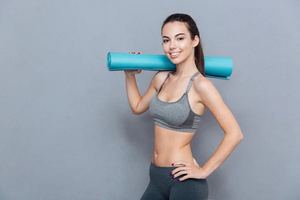 Привлекательная улыбающаяся спортсменка держит коврик для йоги изолированным на сером фоне — стоковое фото