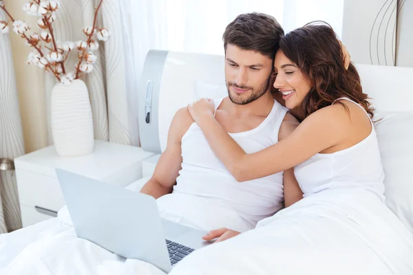 Пара обіймає і використовує комп'ютер, що лежить на їхньому ліжку — стокове фото
