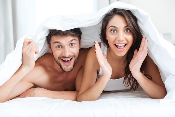 Feliz casal sorridente deitado na cama coberto com cobertor — Fotografia de Stock