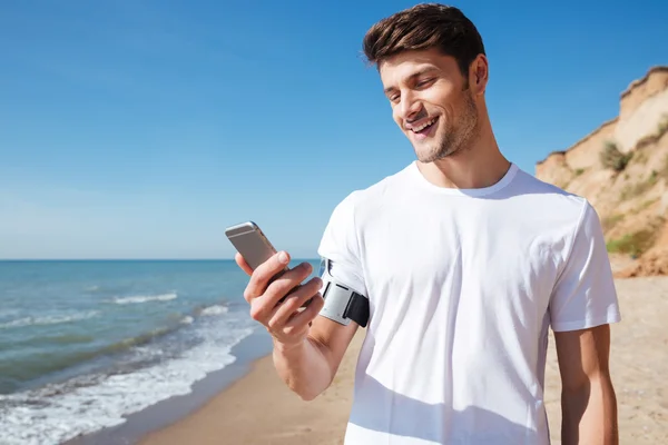 Giovane sportivo allegro che utilizza lo smartphone sulla spiaggia — Foto Stock