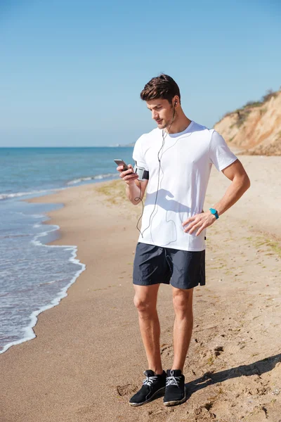 Αθλητικός τύπος ακούτε μουσική χρησιμοποιώντας το κινητό στην παραλία — Φωτογραφία Αρχείου