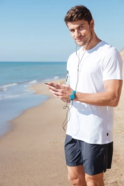 นักกีฬาที่ยิ้มฟังเพลงจากสมาร์ทโฟนบนชายหาด — ภาพถ่ายสต็อก