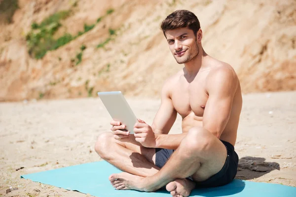Hombre sonriente sentado y usando la tableta en la playa —  Fotos de Stock
