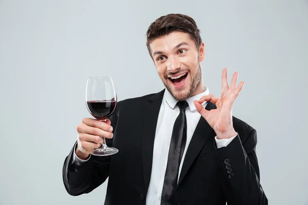 Веселый пьяный бизнесмен пьет красное вино и показывает знак ОК — стоковое фото