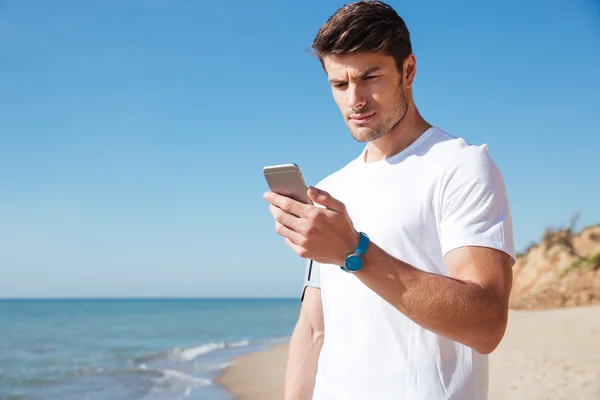 Αθλητικός τύπος στέκεται και χρησιμοποιώντας κινητό τηλέφωνο στην παραλία — Φωτογραφία Αρχείου