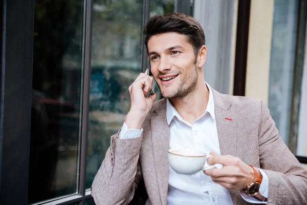 Усміхнений молодий бізнесмен розмовляє на мобільному телефоні у відкритому кафе — стокове фото