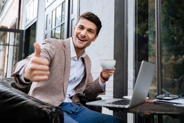 Homme joyeux avec ordinateur portable montrant pouces vers le haut dans un café extérieur — Photo
