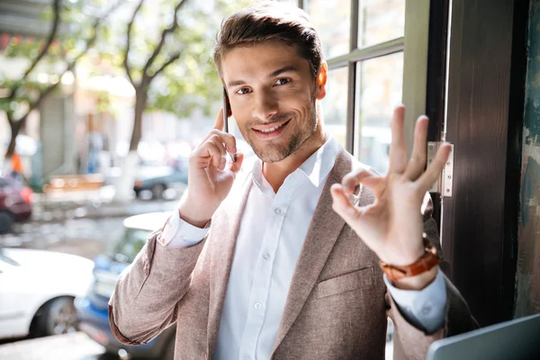 Businessman holding mobile phone and showing ok gesture indoors — Stock Photo, Image