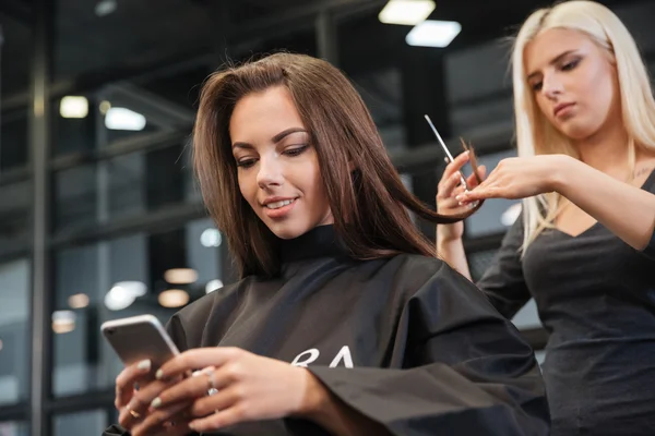 Frau mit Smartphone und Friseur frisiert im Salon — Stockfoto