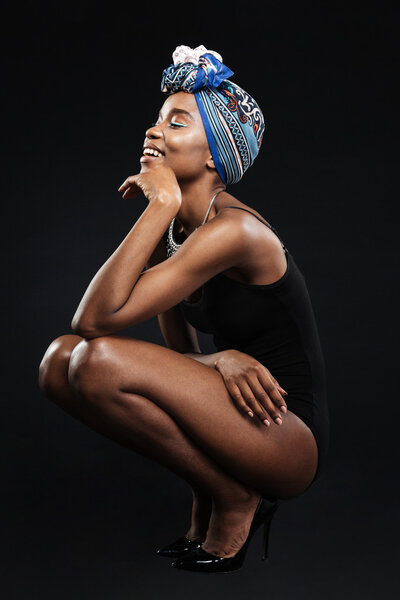Beautiful smiling african woman in bodysuit posing in studio sitting isolated on the black background