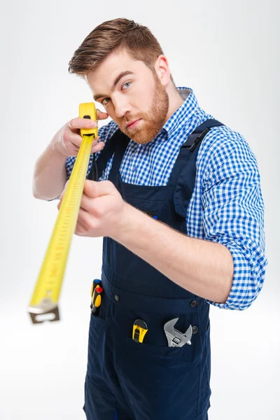 Concentrated bearded young worker in overalls using measuring tape — Stock Photo, Image