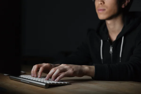 Серьезный азиатский молодой человек, печатающий на клавиатуре в темной комнате — стоковое фото