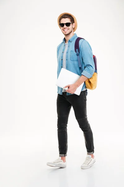 Fröhlicher junger Mann mit Rucksack zu Fuß und Laptop in der Hand — Stockfoto