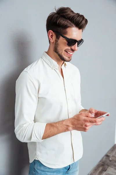 Feliz joven atractivo en gafas de sol de pie y el uso de teléfono inteligente — Foto de Stock