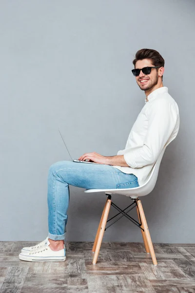 Веселый человек в солнечных очках сидит на стуле и использует ноутбук — стоковое фото
