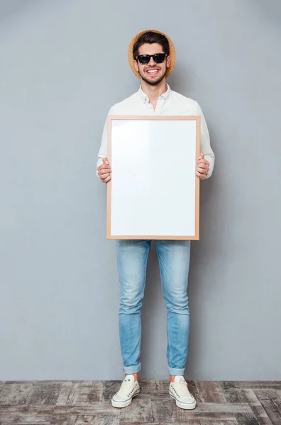 Homme heureux dans des lunettes de soleil debout et tenant tableau blanc vierge — Photo