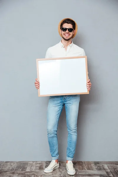 Jeune homme souriant en chapeau et lunettes de soleil tenant tableau blanc — Photo