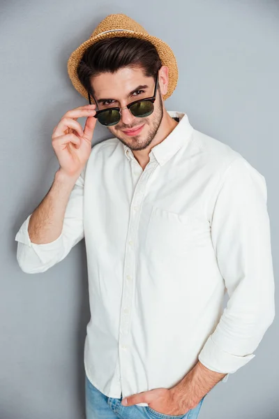 Sonriente joven con sombrero de pie y mirando por encima de las gafas de sol — Foto de Stock