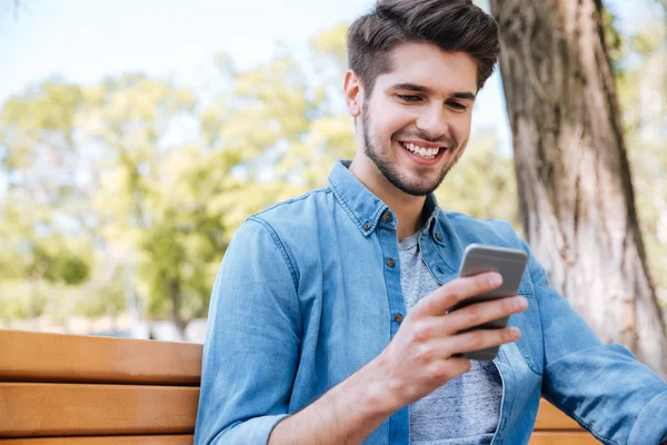 Joven sonriente usando un teléfono inteligente sentado en el banco al aire libre — Foto de Stock