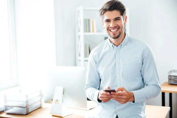 Χαμογελώντας επιχειρηματίας στέκεται και η χρήση του κινητού τηλεφώνου στο γραφείο — Φωτογραφία Αρχείου