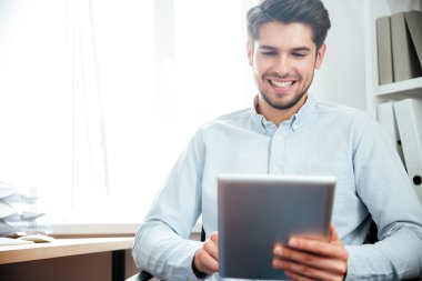 Gülümseyen işadamı otururken ve ofiste tablet bilgisayar kullanarak