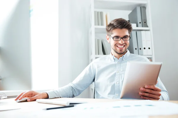 Улыбающийся бизнесмен сидит и пользуется планшетным компьютером в офисе — стоковое фото