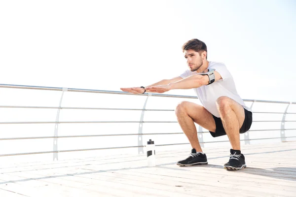 Young handsome sportsman doing squatting outside at the pier — Stock Photo, Image