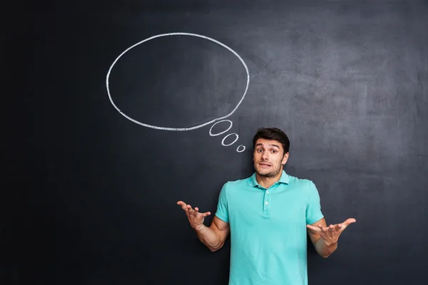 Confused man shrugging shoulders with thinking bubble on blackboard background — Stock Photo, Image
