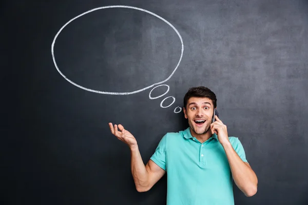 Surprised young man talking on mobile phone over blackboard background — Stock Photo, Image