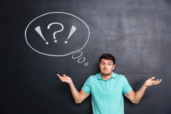 Confused perplexed young man shrugging shoulders over chalkboard background — Stock Photo, Image